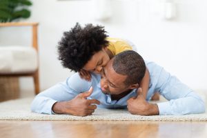 Joyful Kid boy in yellow casual kissing and cuddling his Dad in blue shirt on wood floor in room. Happy smiling African Father and little child son playing at home together. Black family and christian counseling in Atlanta, GA with male counselor William Hemphill for Online therapy in Georgia.