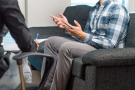 Photo of a man with hands up in a counseling session representing an anxious professional who struggles with Imposter Syndrome and is talking about it with his therapist.