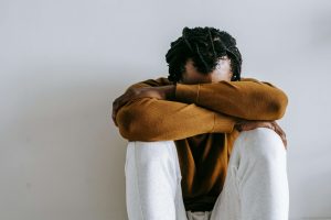 A Black man sitting on the floor with his head resting on his arms, wearing a brown sweatshirt and white pants. This image represents the emotional weight of self-doubt and the impact of relational pain. Counseling services like premarital counseling in Atlanta, GA, and affair recovery in Atlanta, GA can support individuals working through shame, disconnection, and healing after betrayal.