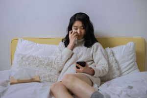 Young woman sitting on a bed, holding a tissue to her eye and looking down at her phone, appearing distressed. Perfect for a blog on affair recovery in Atlanta, GA, and EMDR therapy in Atlanta to heal from betrayal and feelings of inadequacy.