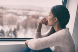 A woman gazes out a window with a thoughtful, distant expression, capturing loneliness during the holidays. Fits content about online marriage counseling in atlanta, ga, marriage counseling online in atlanta, ga, and working with a marriage counselor in atlanta, ga.