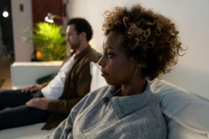 Black woman sitting on couch looking distant with partner in background, showing emotional disconnection from church hurt that decatur marriage counseling and a marriage counselor in atlanta, ga can help couples address and heal.