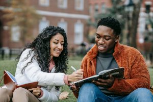 Two smiling Black college students sitting on campus lawn studying together with notebooks, representing the positive connections and support systems that help manage stress, similar to the guidance offered through Black marriage counseling in Atlanta, GA and Decatur marriage counseling