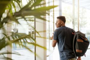 A Black male college student with backpack standing alone looking out a window in a campus building, representing the isolation and stress students experience that can be addressed through support from a Black therapist Decatur GA offering EMDR therapy in Atlanta