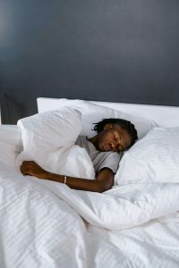 Young Black person resting in bed with white pillows and sheets, symbolizing the peace and better sleep that college students can achieve through anxiety treatment Decatur with a therapist for Black men Decatur