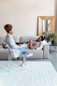 A Black man relaxes on a gray couch during a therapy session while a female therapist sits across from him taking notes, representing the safe and supportive space offered through therapy for black men decatur, ga with a christian therapist in decatur, ga.