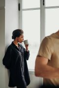 A woman in a black shirt drinking from a mug while looking away thoughtfully by a window, with another person blurred in the foreground. Represents the emotional distance and processing that happens during spiritual trauma therapy in atlanta, ga with a christian therapist atlanta.