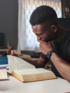 A Black man wearing glasses sits in deep thought with his hand to his face while reading an open Bible at a table, representing the spiritual struggle and questions that can be addressed through anxiety treatment decatur with a black christian therapist atlanta.