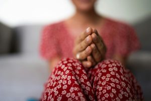 A woman in a coral shirt and red floral pants sits with her hands clasped in prayer position, representing the struggle between faith and pain that accompanies spiritual trauma and can be healed with support from a christian trauma therapist atlanta.
