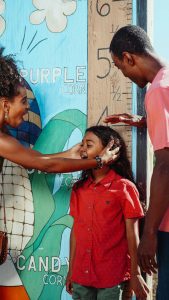 A Black family with two parents and their young daughter in a red shirt smile and measure her height against a colorful mural, representing the joy and connection that can be restored through black marriage counseling in atlanta, ga and support from a therapist for black men decatur.
