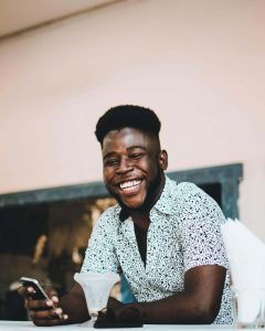 A young Black man smiles broadly while holding his phone at a table, representing the joy and confidence that becomes possible through working with a therapist for black men decatur and receiving anxiety treatment decatur at Faith and Family Empowerment.