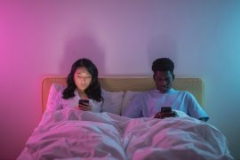 An interracial couple lies in bed in a darkened room each scrolling separately on their phones, representing the parallel living and emotional disconnection that marriage counseling in decatur, ga and a marriage counselor decatur ga at Faith and Family Empowerment can help couples move beyond.