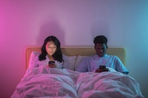 An interracial couple lies in bed in a darkened room each scrolling separately on their phones, representing the parallel living and emotional disconnection that marriage counseling in decatur, ga and a marriage counselor decatur ga at Faith and Family Empowerment can help couples move beyond.