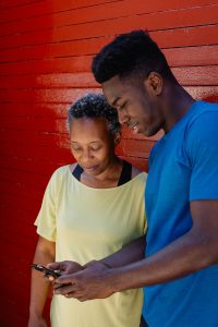 An older Black woman and a young Black man lean together against a red wall smiling while looking at a phone, representing the family connection and generational bonds that christian counseling in decatur, ga and anxiety treatment decatur at Faith and Family Empowerment can help families protect and strengthen.