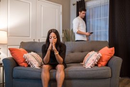 A woman sits on a gray couch with her hands clasped to her face while her partner stands in the background scrolling his phone, representing the emotional distance and disconnection that marriage counseling in decatur, ga can help couples work through.
