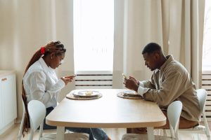 A Black couple sits across from each other at a dining table during a meal, both absorbed in their phones without connecting, representing the silent disconnection that premarital counseling in atlanta, ga can help prevent and affair counseling atlanta at Faith and Family Empowerment can help couples heal from.