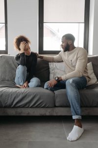 A Black man reaches out to comfort his distressed partner on a gray couch, representing the difficult but necessary conversations that premarital counseling in atlanta, ga and therapy for black men decatur, ga at Faith and Family Empowerment can help couples navigate.