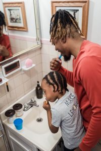 A Black father and child brush their teeth together in the morning, representing the daily routines that consume couples during busy seasons and the support available through affair counseling atlanta and a black therapist decatur ga.