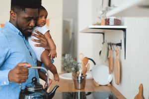 A Black father holds his baby while multitasking in the kitchen, representing the daily demands of parenthood that online therapy in georgia and a black therapist decatur ga at Faith and Family Empowerment can help couples navigate.