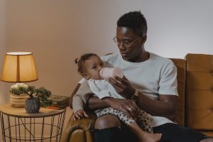 Feeding the Baby | couples counseling decatur | online therapy in georgia | marriage intensive decatur | marriage counselor decatur ga | Sandy Springs, GA | Brookhaven, GA | Decatur, GA A Black father bottle feeds his baby in a cozy armchair, representing the intentional support that christian marriage counseling in decatur, ga and couples counseling decatur can help couples build.